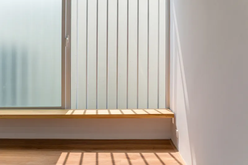Detail of timber window sill with vertical metal screen casting shadow stripes across wood flooring