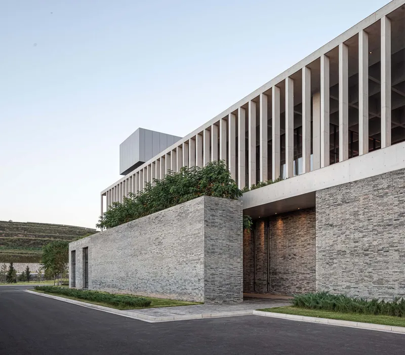 Layered stone entrance wall with recessed doorway beneath the projecting slatted timber volume and climbing greenery