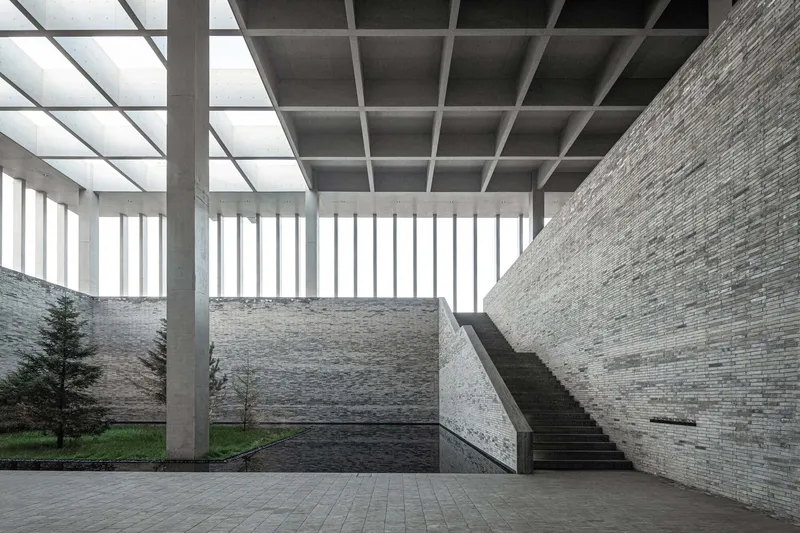 Interior courtyard with stone walls, concrete columns supporting a coffered skylight, and a staircase under diffused light