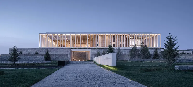 Illuminated vertical louver facade atop a stone plinth with a gravel entry path at dusk