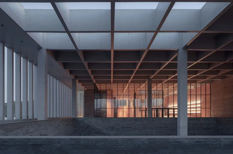 Covered colonnade with concrete columns and timber grid ceiling overlooking a reflecting pool at dusk