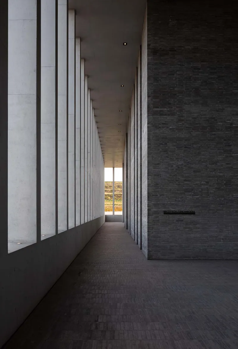Long corridor with rhythmic vertical concrete fins casting shadows on dark brick flooring at dusk