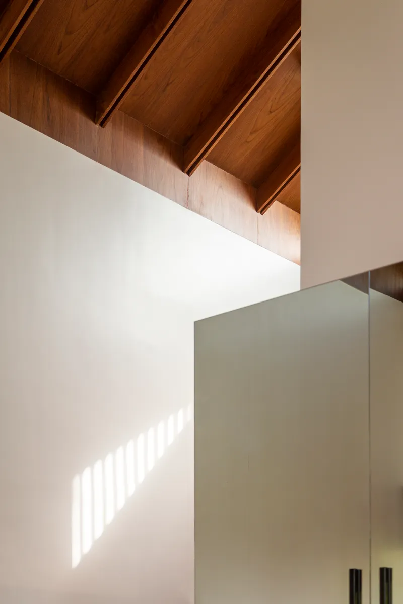 Double-height corner detail showing timber ceiling beams and sunlight patterning a white wall