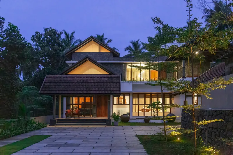Front elevation showing gabled pavilion, paved courtyard, and illuminated interiors amid palm trees at dusk