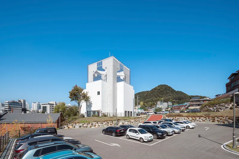 White tower standing above a parking lot with hillside and scattered trees visible in the background