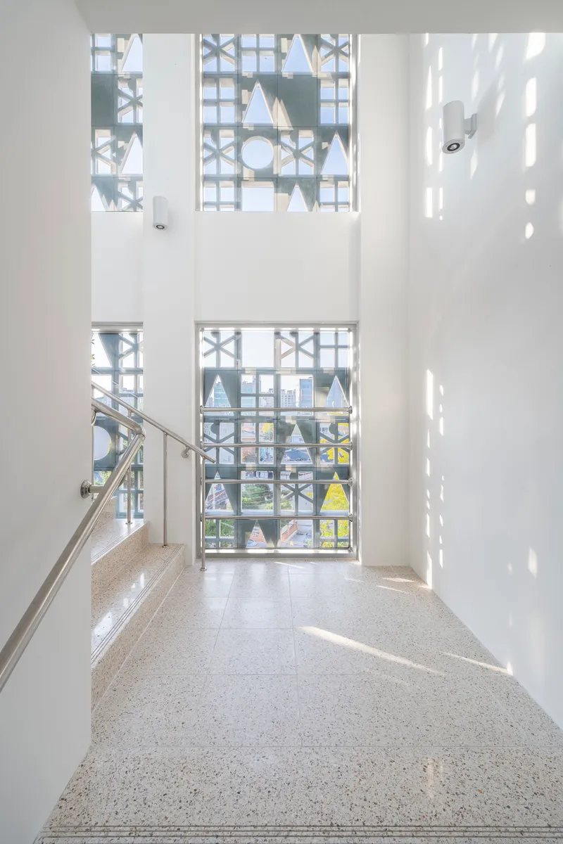 Double-height stairwell with terrazzo flooring and geometric window screens filtering bright daylight patterns