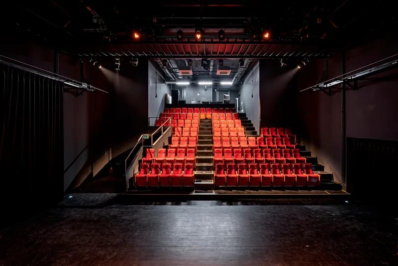 View from the stage toward the tiered red seating under exposed black ceiling trusses and lighting rigs
