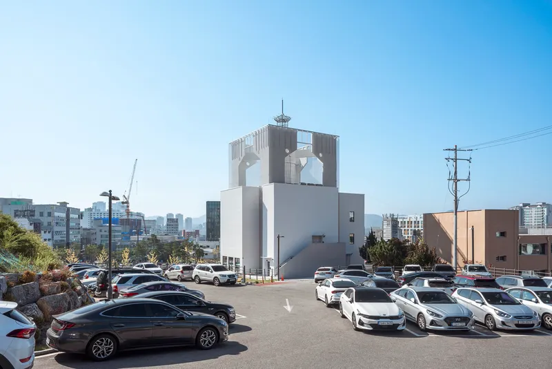 White tower with arched openings rising above a parking lot filled with cars under clear blue sky