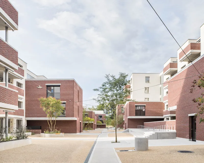 Shared courtyard with red brick housing blocks, young trees, and concrete seating under cloudy skies