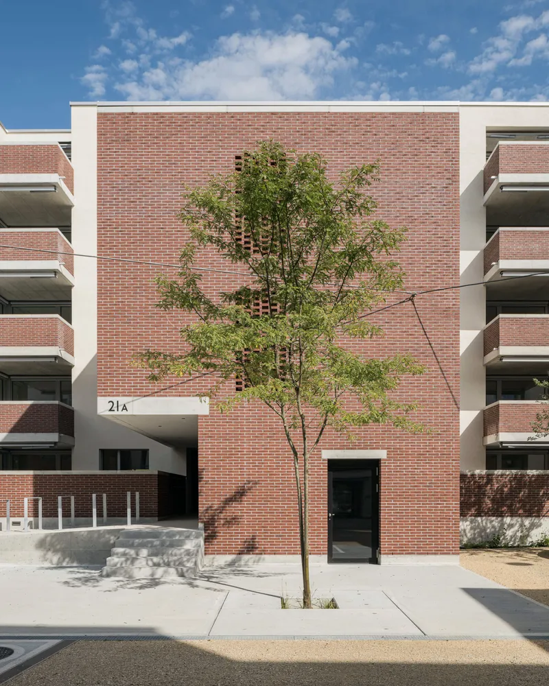 Red brick facade with recessed entry, white balconies flanking, and young tree in foreground