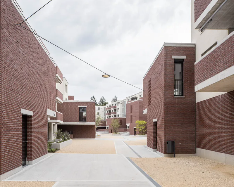 Pedestrian lane between red brick volumes with gravel paving and overhead utility lines