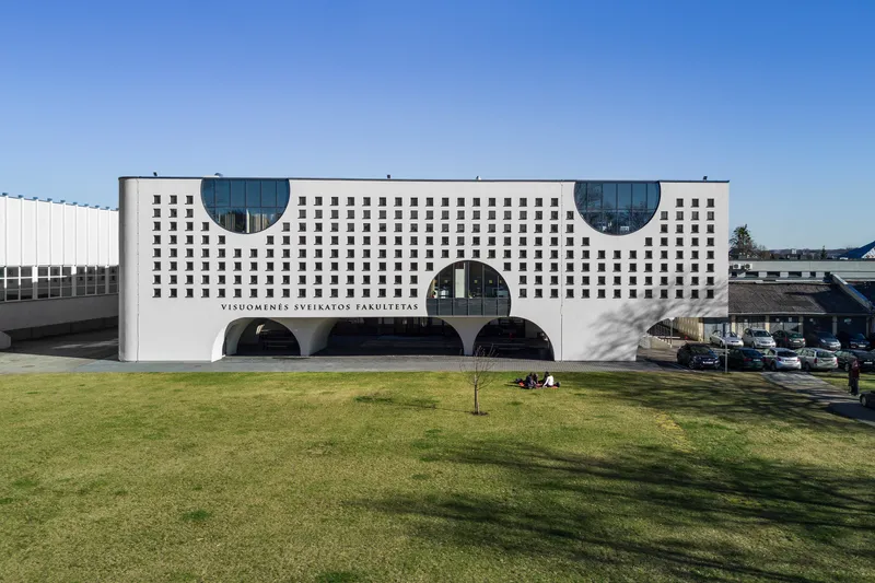 White perforated facade with circular glazed cutouts and arched openings at ground level across a green lawn