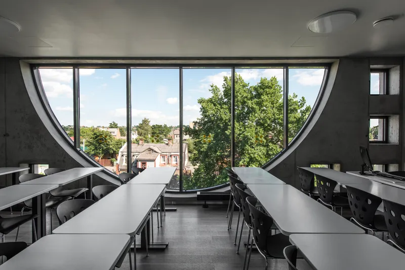 Interior classroom with rows of tables facing a semicircular window overlooking leafy trees and rooftops
