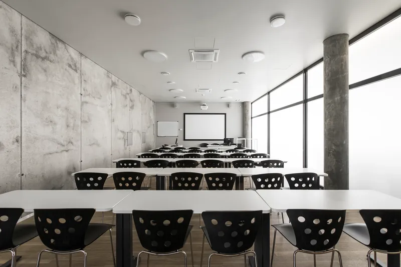 Classroom with rows of white tables and black perforated chairs facing projection screen