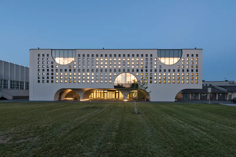 Front elevation showing the gridded white facade with illuminated arched entrance portals at dusk
