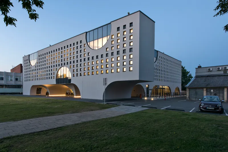 Street view of the white perforated facade with lit arched entrances beneath trees at twilight