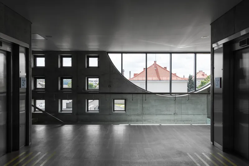 Interior lobby with curved window wall framing red tile roofs and square openings in dark concrete