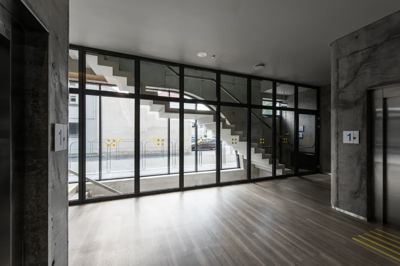 Lobby entrance with black-framed glazing overlooking white staircase and exposed concrete columns