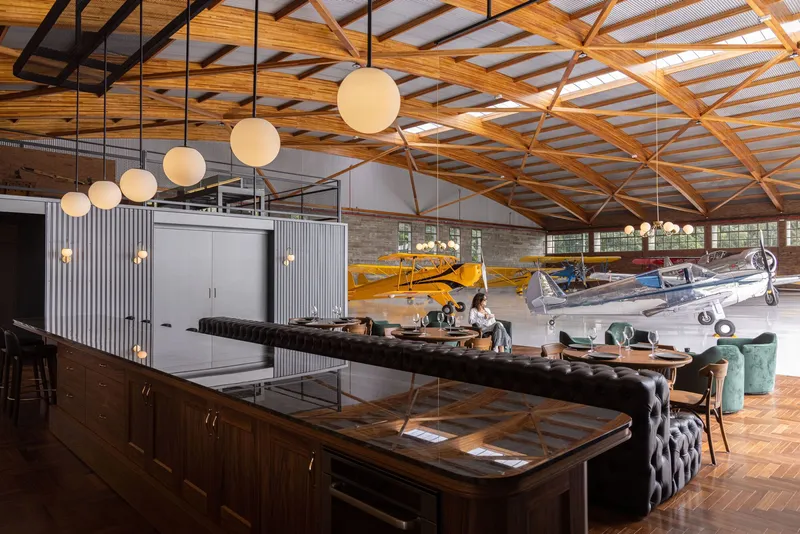 Hangar interior with exposed timber trusses, corrugated metal ceiling and vintage aircraft parked beside a lounge area