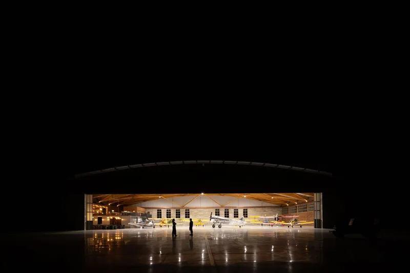 Aircraft hangar interior with exposed timber trusses and parked planes viewed through the open door at night