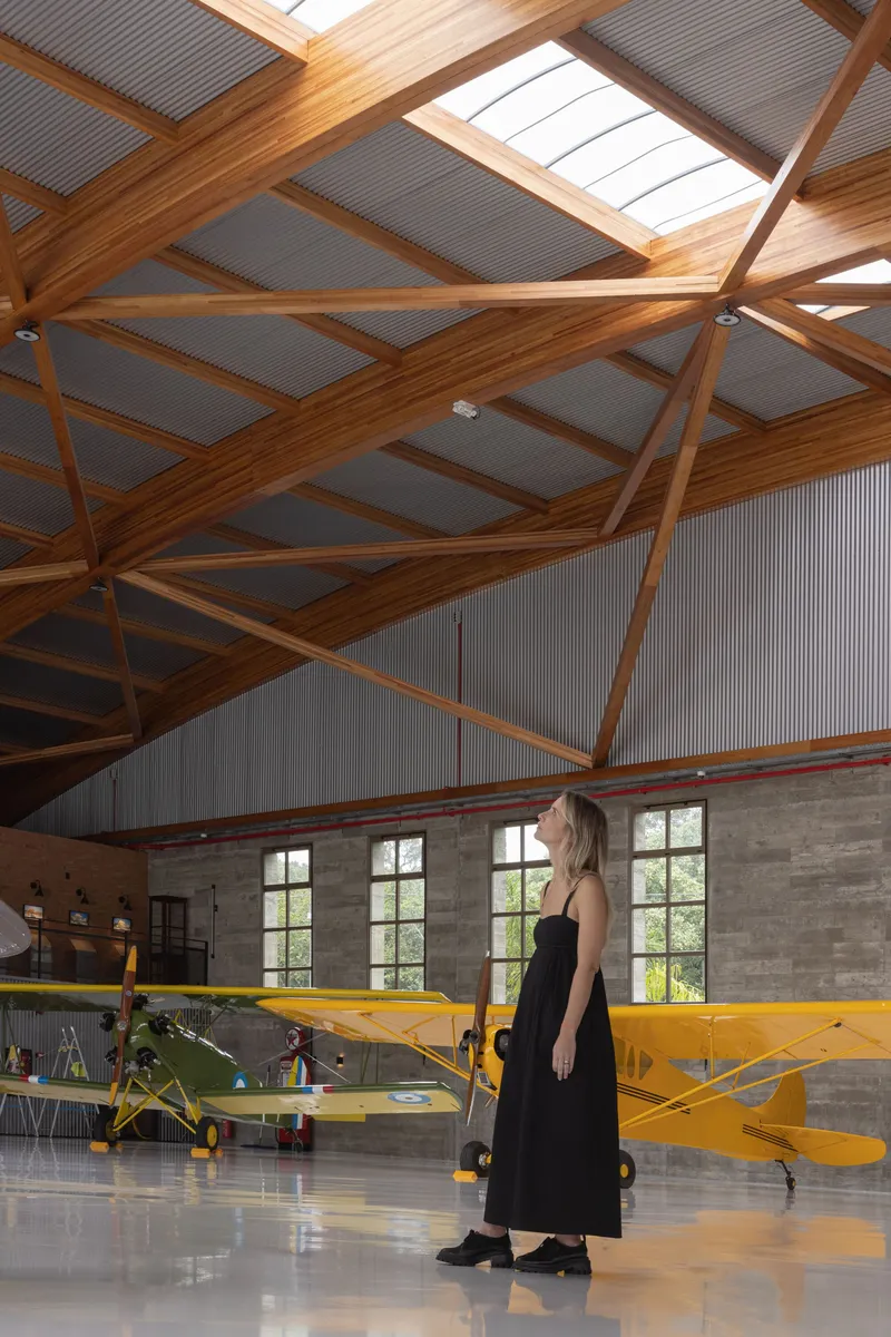 Visitor standing before yellow biplane under the diagonal timber truss ceiling with clerestory glazing