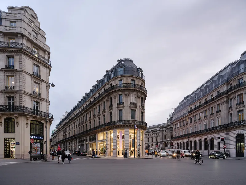 Corner retail building with illuminated ground floor windows at dusk surrounded by pedestrians and cyclists