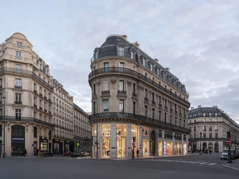Curved masonry corner tower with mansard roof and illuminated ground floor retail space under cloudy sky