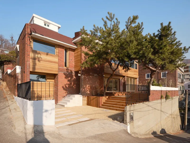 Street-level view of the red brick facade with timber-clad volumes and a mature pine tree in the forecourt