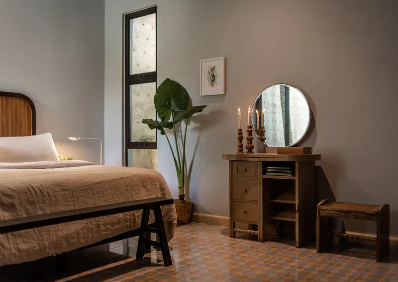 Bedroom corner with tall vertical window, potted bird-of-paradise, and candlelit dresser with round mirror
