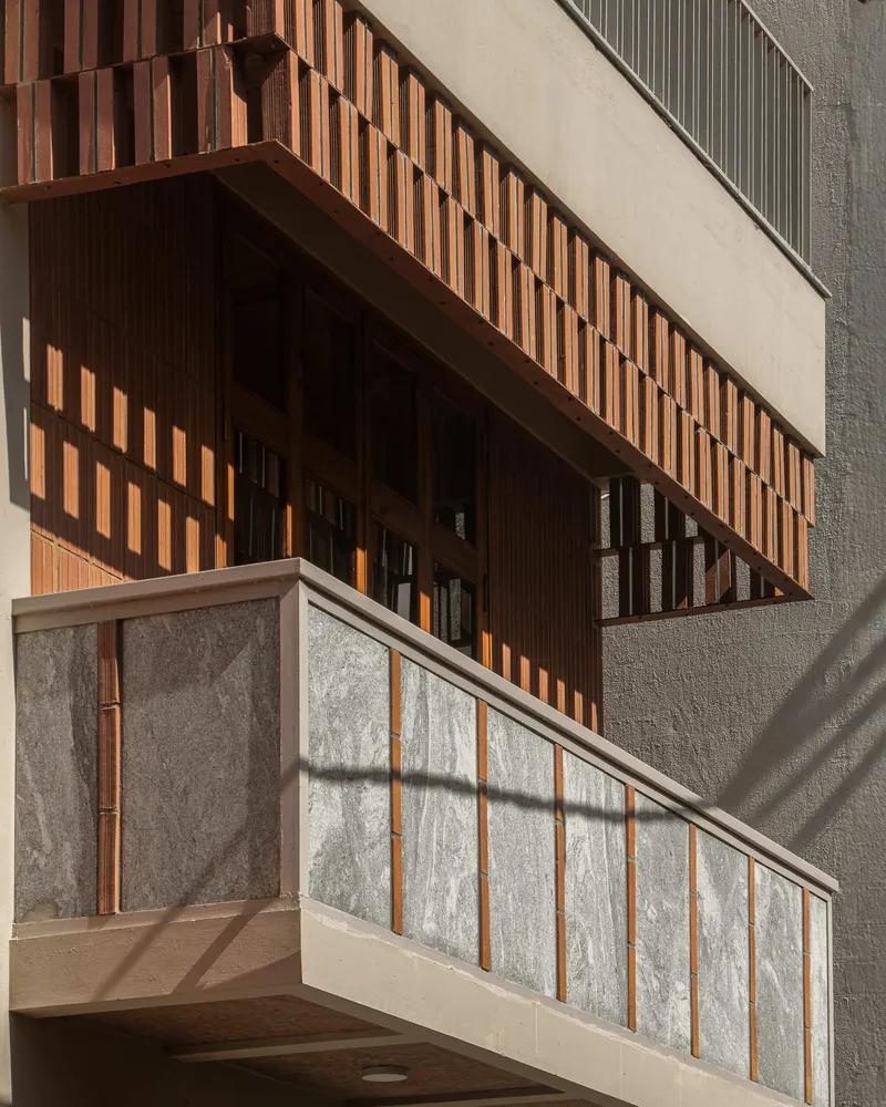 Facade detail showing timber slat balcony screen above textured stone panel railing in afternoon sunlight