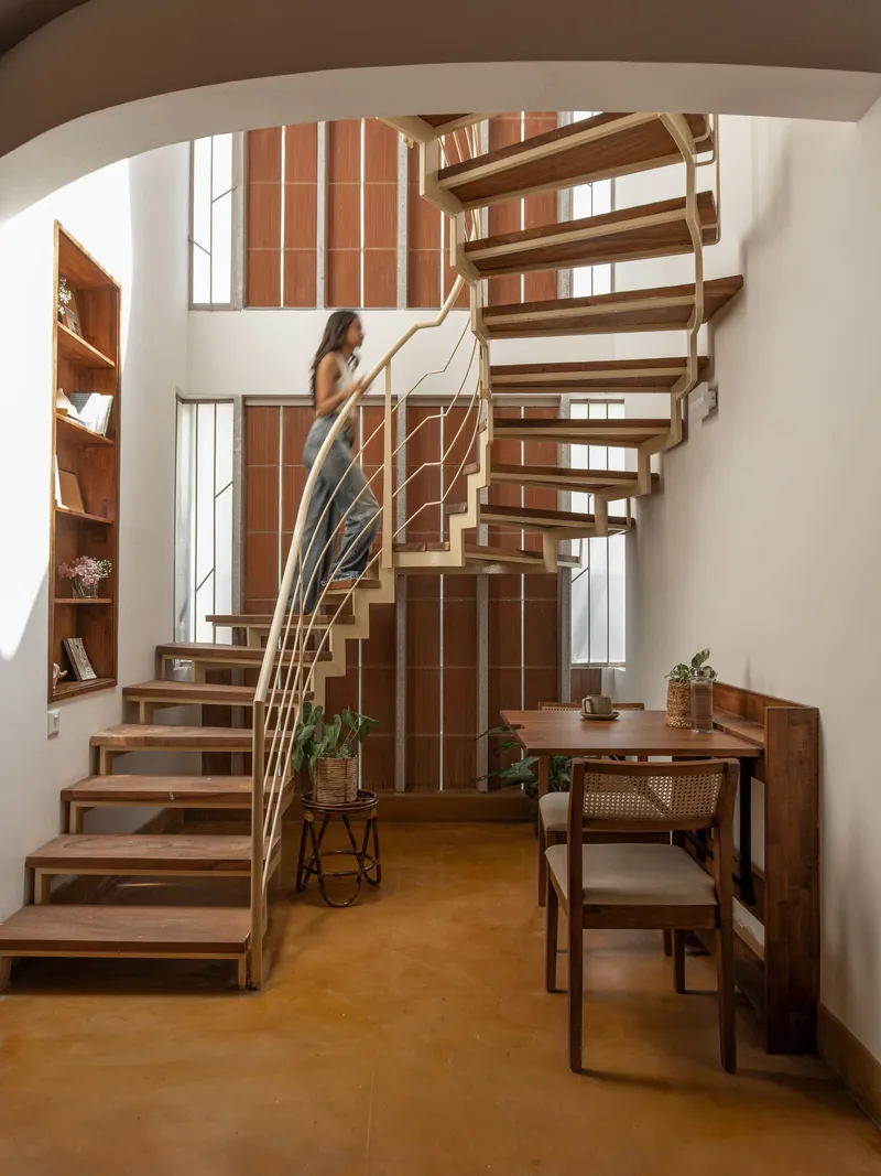 Open-tread timber staircase with curved rope railings rising past a brick wall with vertical windows