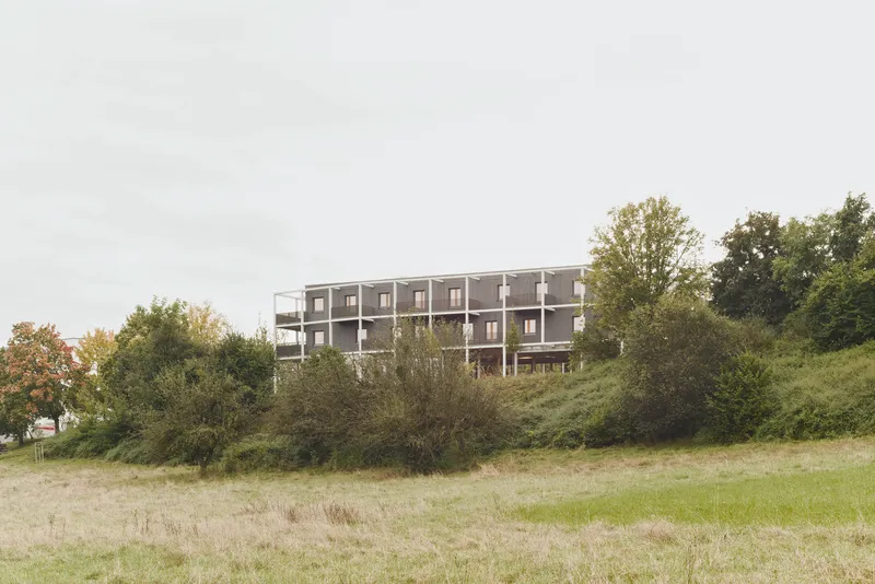 Three-story residential building with white frame and dark cladding on a hillside overlooking a grassy meadow