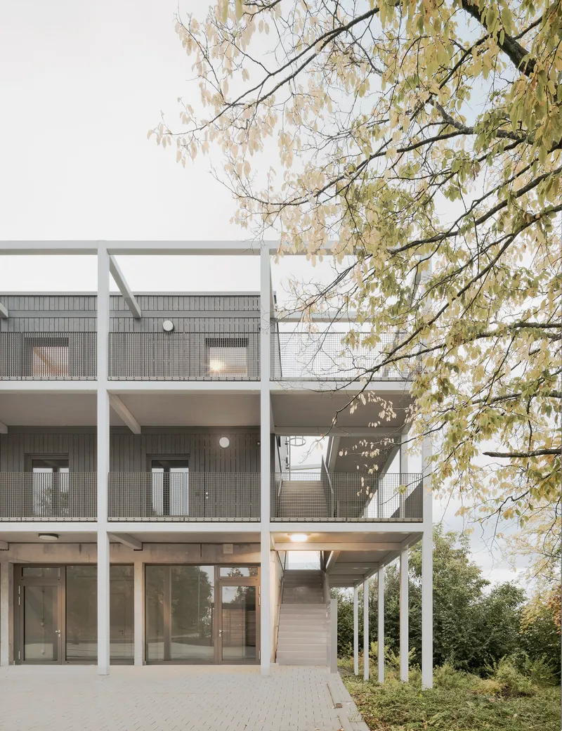 Facade detail showing white structural frame with perforated metal balcony screens beneath overhanging tree branches