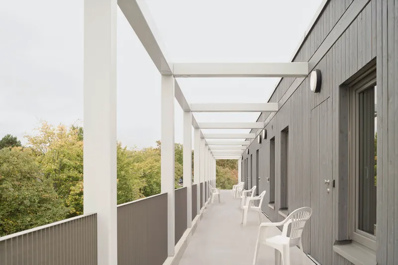 Exterior walkway with white steel pergola, grey cladding and white plastic chairs overlooking green trees