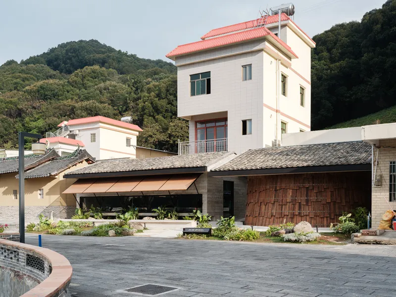 Low-slung pavilion with timber-shingled roof and planted courtyard beside paved plaza and hillside housing