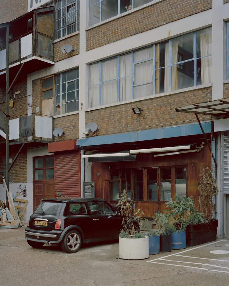 Facade detail showing stacked residential units with balconies and a ground floor shopfront behind planters and a parked car