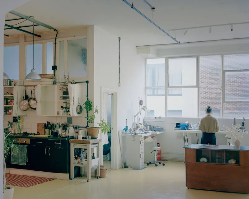 Open-plan studio interior with kitchenette, large windows, and a figure working at a table near the glass