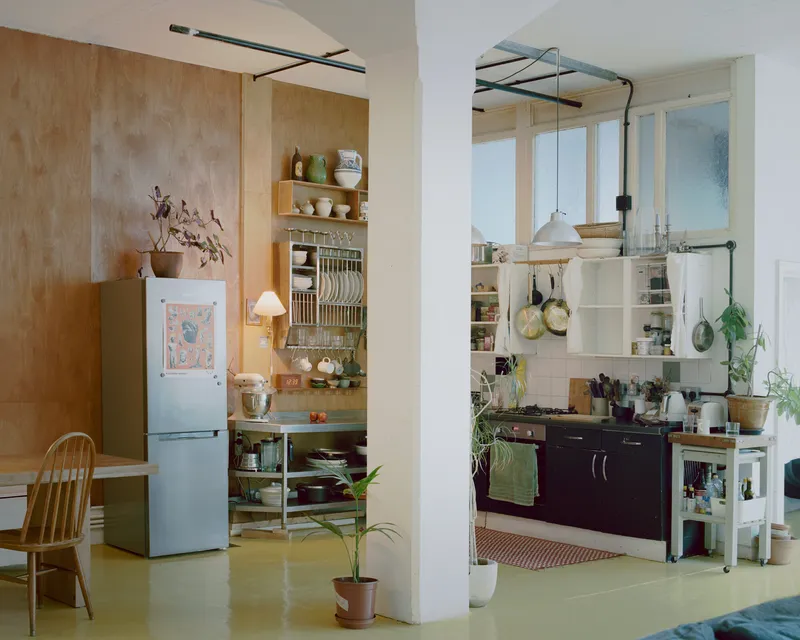 Kitchen alcove with open shelving and dish rack beside plywood partition and exposed piping