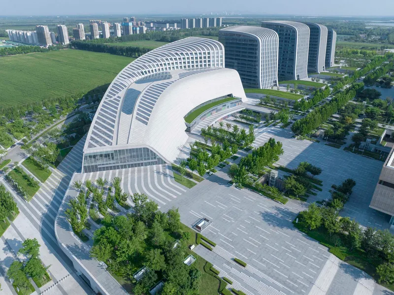 Curved building with horizontal white louvers wrapping a courtyard and entrance plaza under a clear blue sky