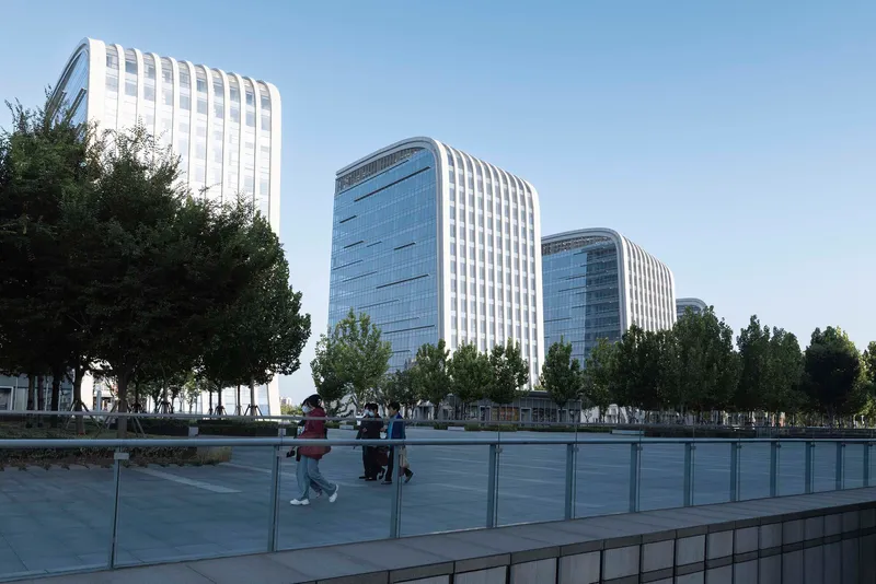 Glass balustrade reflecting pedestrians crossing the paved plaza between the towers and trees
