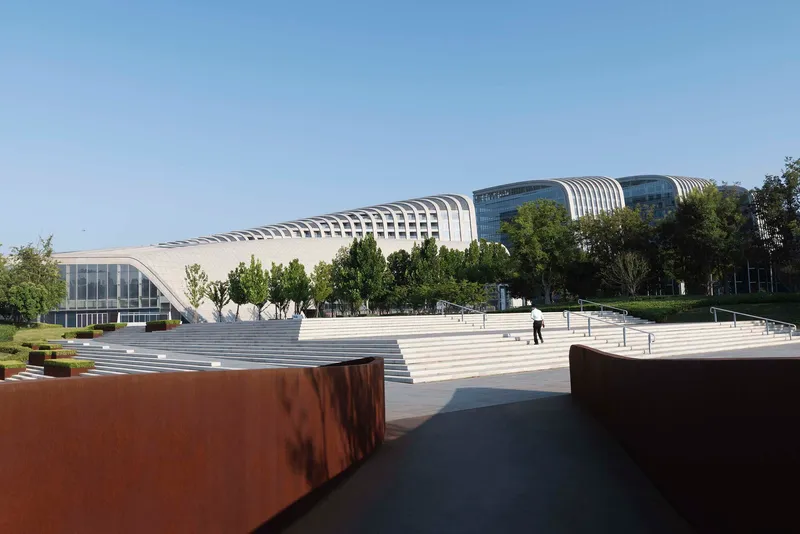Wide plaza with weathered steel walls and white stepped terracing leading to the ribbed barrel-vault facade