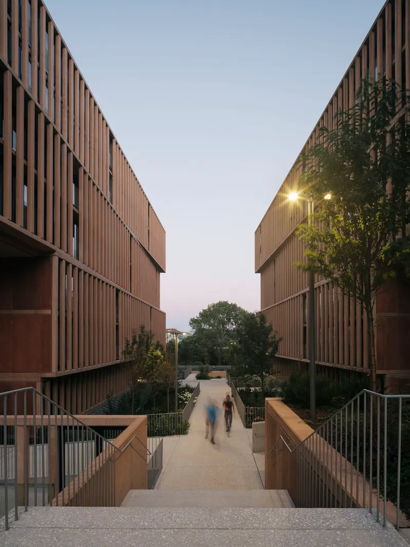 Pedestrian bridge between two timber-clad buildings with residents walking at dusk