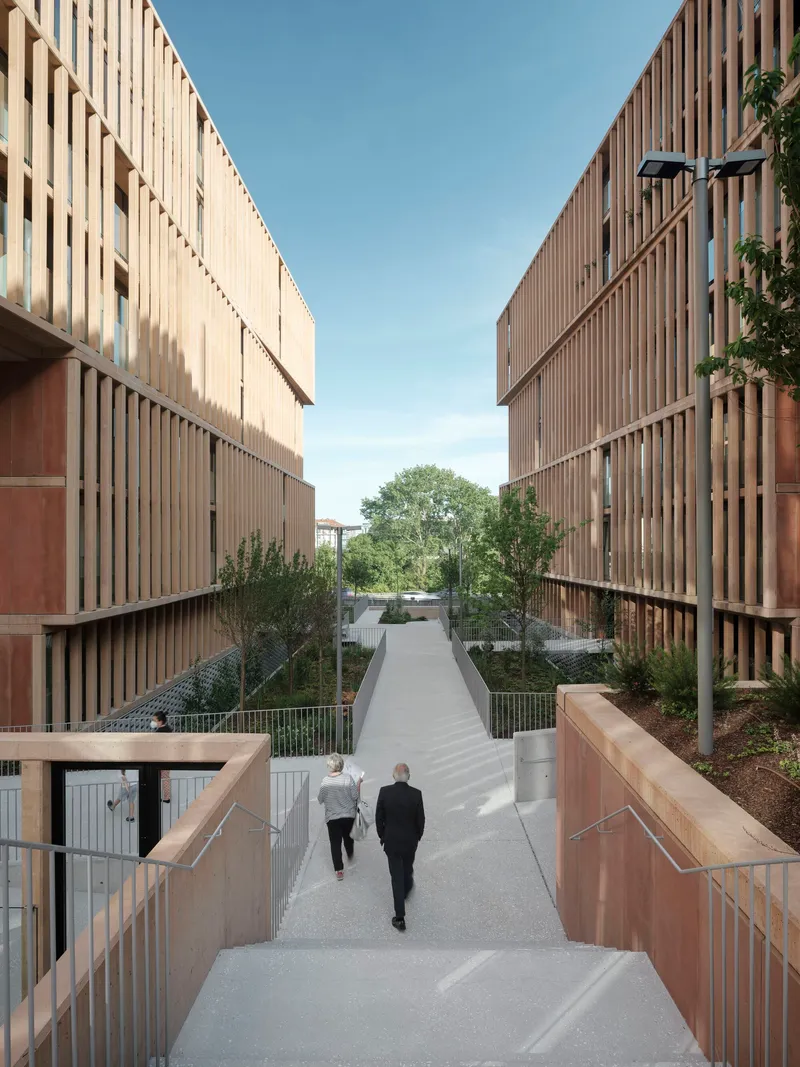 Pedestrian pathway between vertical timber-screened facades with two people descending the sloped walkway