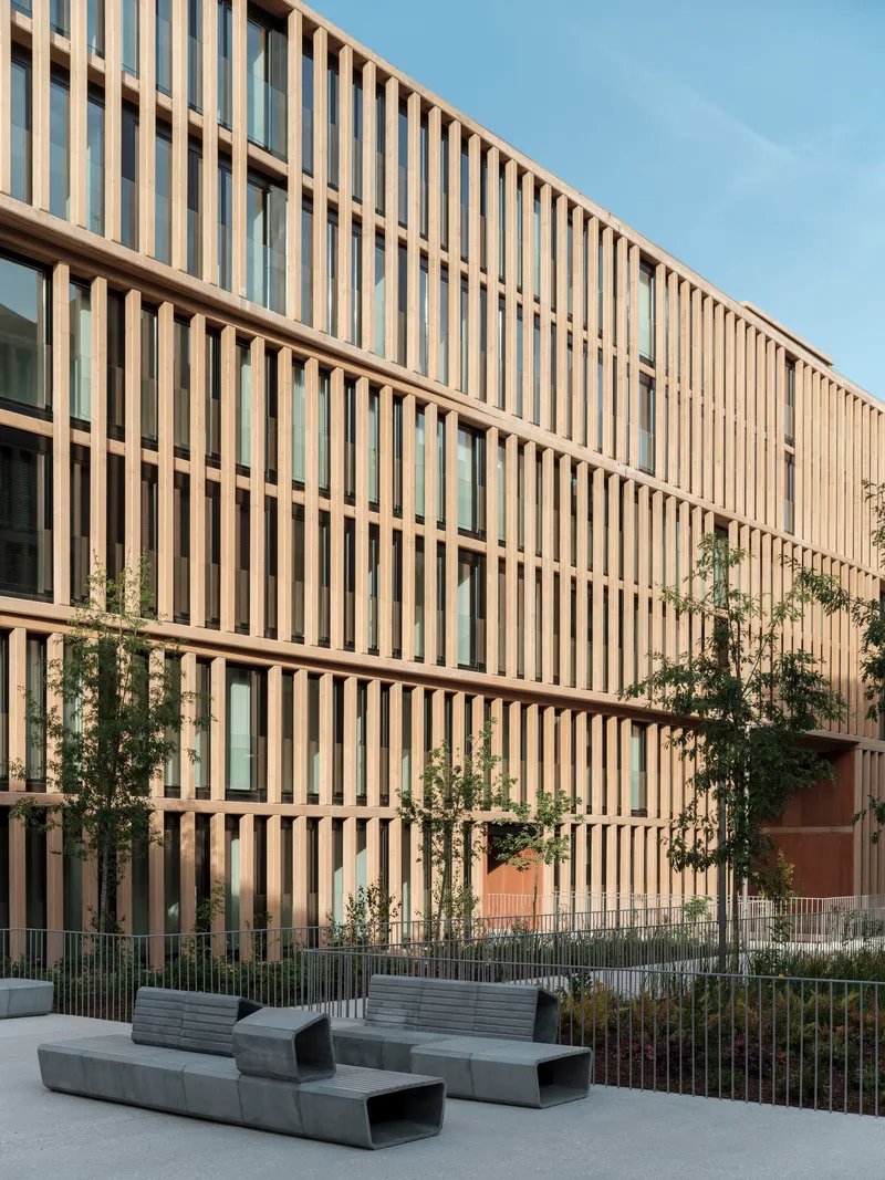 Vertical timber brise-soleil facade with concrete benches and young trees in the courtyard below