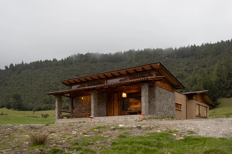 Front facade with stone columns and deep overhangs set on gravel site beneath low fog and forested slopes