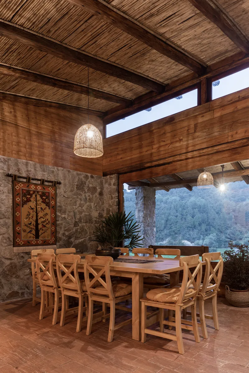 Open dining pavilion with woven pendant lamps and mountain view through timber columns at dusk