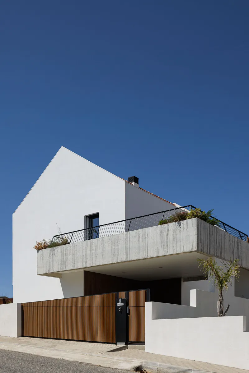 White gabled volume with projecting concrete balcony, vertical timber gate and palm beside the street