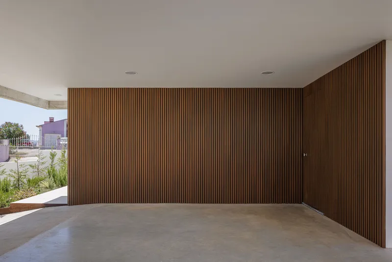 Covered carport with vertical timber slat wall and polished concrete floor opening to planted surroundings