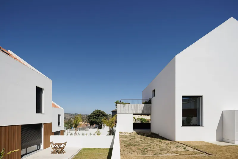 Courtyard between white gabled volumes with a planted lawn and wooden table under clear sky