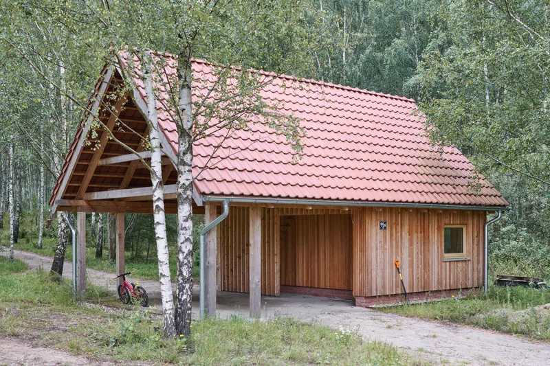 Timber cabin with red tile roof and open carport framed by birch trees on a gravel clearing